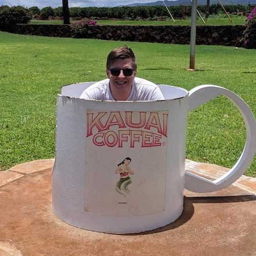 A picture of me, a white man with short light brown hair crouching down in a giant metal coffee cup that has "Kauai Coffee" written on the side and a logo of a women in a hula skirt. 